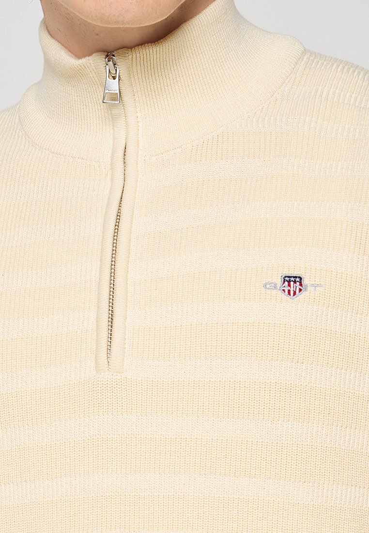 Gros plan sur un pull en tricot de couleur crème avec une fermeture éclair demi-longueur et un petit logo écusson rouge, blanc et bleu sur la poitrine.