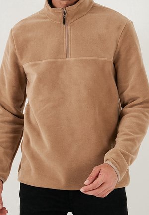 Mężczyzna ubrany w beżową bluzę polarową z przednim zamkiem na pół i długimi rękawami, w zestawieniu z czarnymi spodniami.