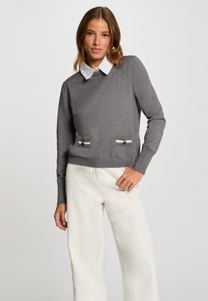 Maglione grigio con maniche lunghe, polsini a coste e due tasche frontali decorate con strisce bianche e bottoni decorativi. Collo bianco.
