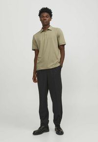 Polo shirt verde oliva con maniche corte, colletto e pattina con tre bottoni, abbinato a pantaloni scuri su misura e scarpe nere.