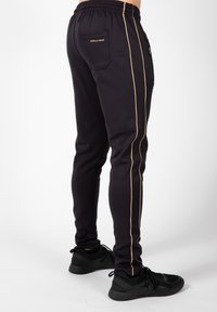 Zwarte sport joggers met gouden zijkanten, elastische tailleband, achterzak en een slim fit ontwerp. Gemaakt met zwarte sneakers.