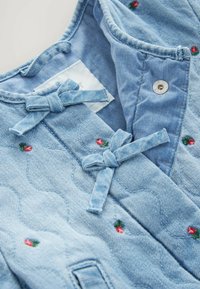 Hellblauer gesteppter Stoff mit kleinen roten Blumenstickereien, gebundenen Schleifen und einem Metall-Druckknopf im Kragenbereich eines Kleidungsstücks.