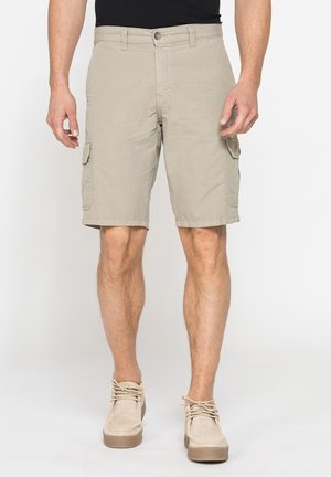 Uomo che indossa pantaloncini cargo beige e scarpe casual beige in piedi davanti a uno sfondo semplice.