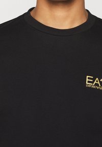 Svart bomulls t-shirt med rund halsringning, prydd med en liten guld Emporio Armani-logotyp på vänster bröst. Slät textur.