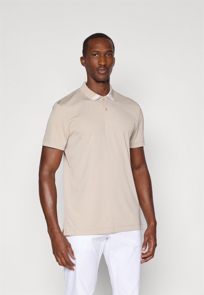 Beige polo skjorte laget av glatt stoff, med en klassisk krage med hvite detaljer og knappestolpe. Brukt med hvite bukser.