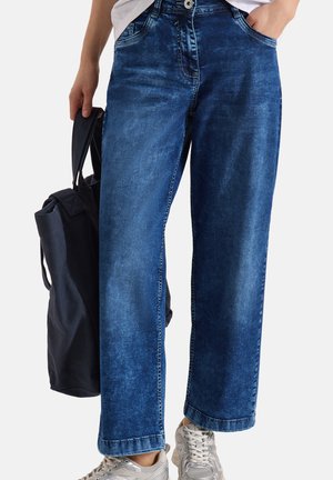 Persona che indossa jeans blu larghi, sneaker bianche, tiene una borsa scura nella mano sinistra, mano destra in tasca.