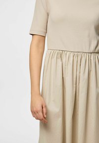 Bež haljina s kratkim rukavima, koja ima uski gornji dio i naboranu, lepršavu suknju. Glatka tekstura s suptilnim sjajem.