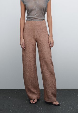Femme portant un pantalon large en lin marron taille haute, un top gris à manches courtes, un collier et des sandales noires, debout devant un fond uni.