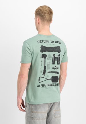 Mann trägt grünes T-Shirt mit schwarzen Survival-Grafiken und Text "Return to Base" und "Alpha Industries" auf dem Rücken.