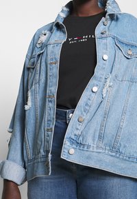 Lichtblauwe denimjacket met versleten details, knoopsluiting en omgevouwen mouwen, gedragen over een zwart T-shirt. De jeans zijn ook donkerblauw.