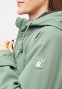 Zelená lehká bunda s vysokým límcem, stahovacími šňůrkami a logem na levém rukávu. Hladká textura, neformální design.