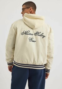 Kremowa bomberka z kapturem, z haftowanym napisem "Nautica Sailing Crew" na plecach oraz paskowanymi ściągaczami przy mankietach i u dołu.