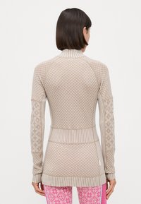 Beige langermet genser med teksturerte mønstre, ribbestrikkede mansjetter og krage. Utformet med en tettsittende silhuett og en buet nederkant.