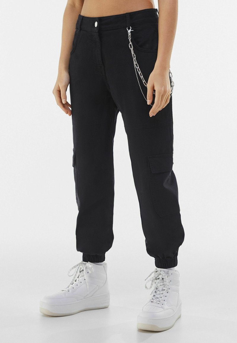 Pantalon cargo noir avec poignets élastiques et détail chaîne argentée porté avec des baskets montantes blanches sur une personne debout.