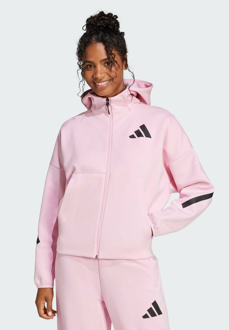 Felpa con zip di colore rosa chiaro, dotata di cappuccio, con strisce nere sui maniche e logo nero sul petto. Tessuto morbido.
