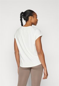 Haut blanc à manches courtes en tissu lisse, col rond, coupe décontractée, associé à un legging beige. La vue de dos montre une silhouette épurée.
