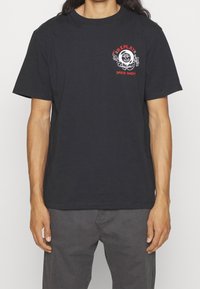 Černé bavlněné tričko se zřetelným červeným a bílým potiskem na hrudi, na kterém je nápis "Replay Speed Shop" s kruhovým designem a plamennými akcenty.