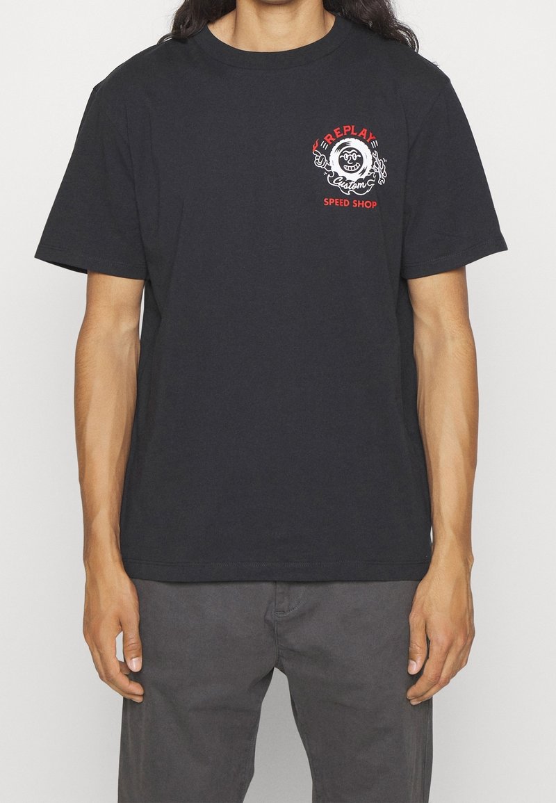 Černé bavlněné tričko se zřetelným červeným a bílým potiskem na hrudi, na kterém je nápis "Replay Speed Shop" s kruhovým designem a plamennými akcenty.