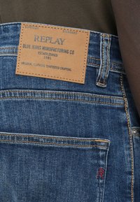 Μπλε τζιν τζιν με καφέ δερμάτινη ετικέτα που φέρει ανάγλυφο κείμενο. Περιλαμβάνει ένα μικρό κόκκινο λογότυπο "R" στην πίσω τσέπη. Τυπικές ραφές λεπτομέρειας.