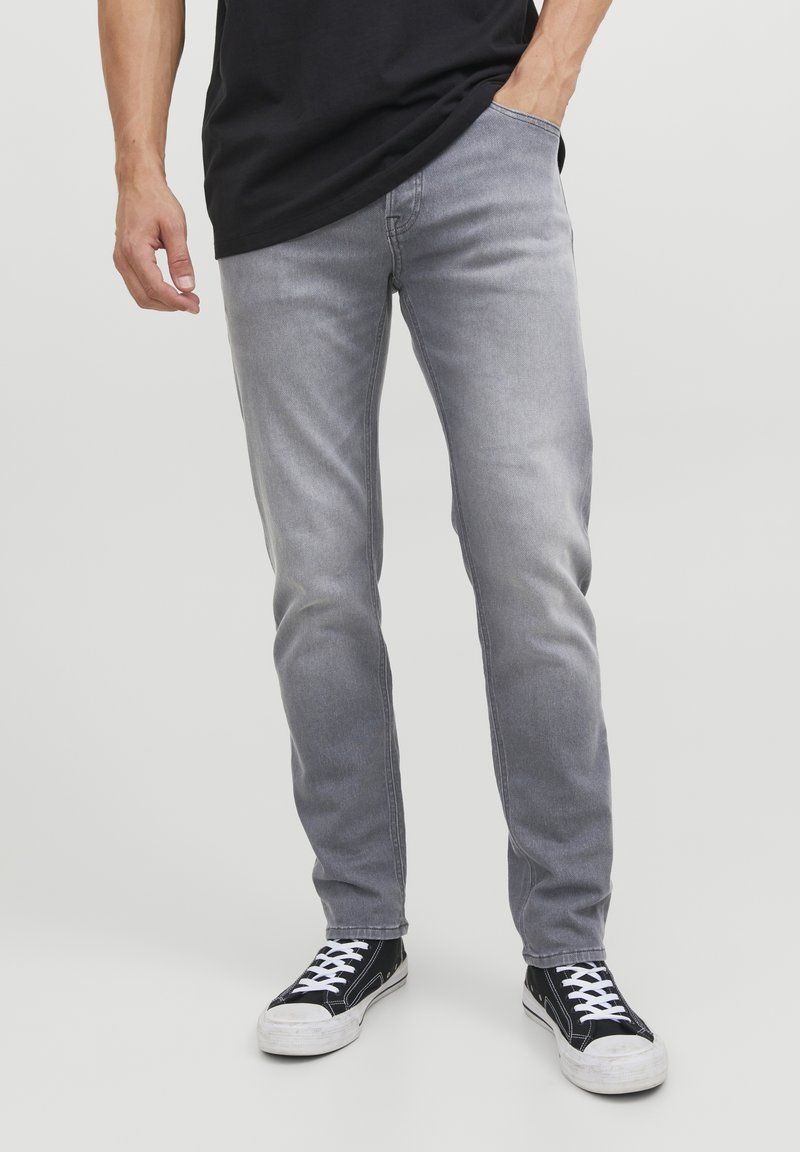 Vaqueros de mezclilla gris con un ajuste slim, que presentan un diseño clásico de cinco bolsillos. Usados con zapatillas negras y una camiseta negra, mostrando una textura sutil.