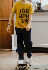 Child wearing yellow shirt and dark pants standing with one foot on a skateboard indoors on a wooden floor.