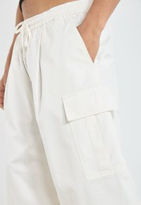 White cotton cargo pants with an elastic waistband, featuring side pockets and a large cargo pocket on the thigh. Smooth texture, relaxed fit.