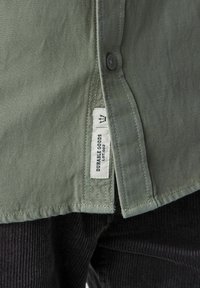 Olivengrünes Hemd mit Knopfleiste aus glattem Material, mit einem Stoffetikett, das an der Seitennaht mit "DURABLE GOODS" versehen ist.