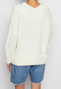 Weißer gestrickter Pullover mit V-Ausschnitt, fallenden Schultern und gerippten Ärmelbündchen, kombiniert mit blauen Denim-Shorts mit ausgefransten Kanten.