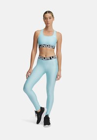 Svetlomodrá športová podprsenka a zodpovedajúce legíny s čiernymi elastickými pásmi s textom „UNDER ARMOUR“. Obuv je čierna s bielou podrážkou.