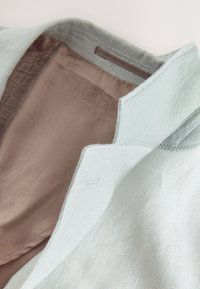 Blazer di lino azzurro chiaro con una fodera interna di raso marrone liscia che presenta un motivo testurizzato sottile. Colletto aperto, cuciture dettagliate visibili.