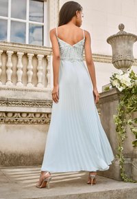 Light blue pleated maxi dress with a fitted, beaded bodice and thin straps. Silver high-heeled sandals visible. Elegant and flowing design.