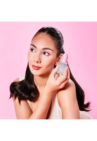 Przezroczysta szklana butelka z różową nakrętką, oznaczona jako „Olejek przeciw puszeniu włosów”. Osoba trzyma ją na różowym tle, prezentując gładkie czarne włosy.