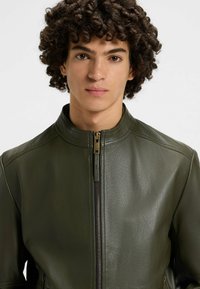 Giacca in pelle verde oliva con colletto alla coreana, caratterizzata da una chiusura frontale con zip e una superficie liscia, che mette in mostra dettagli di design minimalisti.