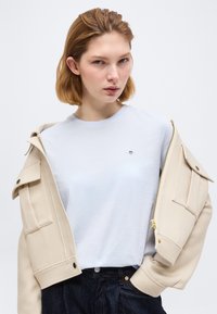 Νεαρή γυναίκα με ανοιχτό καστανά μαλλιά φοράει λευκό t-shirt και μπεζ σακάκι πέταλο στους ώμους, ποζάροντας μπροστά σε μονόχρωμο φόντο.