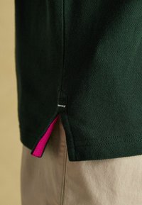 Tumed roheline polo särk tekstureeritud kangast, külgmise lõhiku juures roosa aksent, nähtavad õmblused ja silt hemmi lähedal.