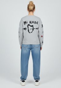 Felpa grigia con testo grafico nero "Mi Amor" e illustrazioni di cuori sulla schiena e sulle maniche. Abbinata a jeans larghi blu chiaro e sneakers.