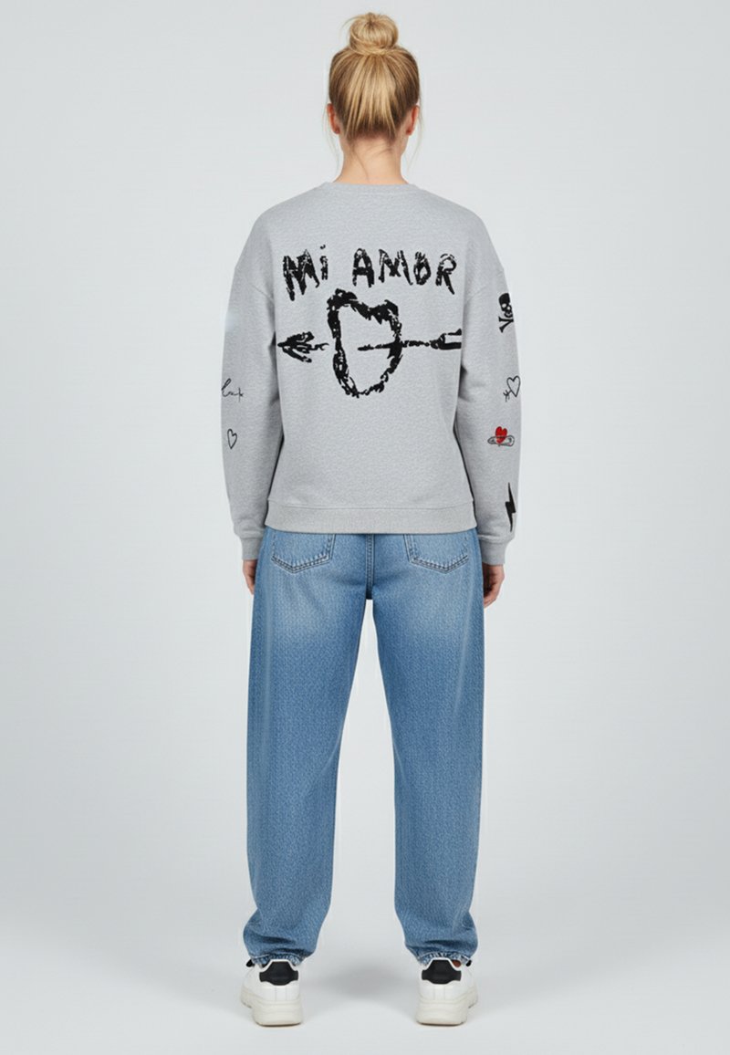 Felpa grigia con testo grafico nero "Mi Amor" e illustrazioni di cuori sulla schiena e sulle maniche. Abbinata a jeans larghi blu chiaro e sneakers.