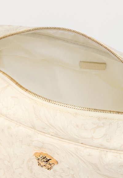 Τσάντα από κρεμ ύφασμα με ανάγλυφο λουλουδένιο σχέδιο, χρυστό φερμουάρ κ αι διακοσμητικό στοιχείο σε σχήμα λιονταριού· το επενδεδυμένο εσωτερικό είναι φωτεινό και απαλό.
