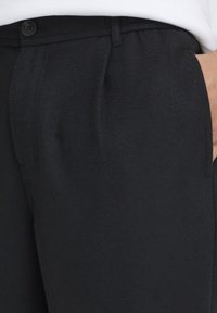 Svarte bukser laget av teksturert stoff, med rett snitt, sidelommer og knappelukking i livet.