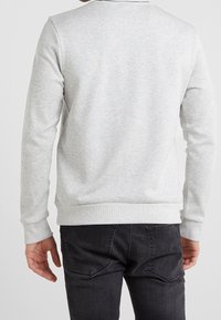 Man med ljusgrå långärmad sweatshirt och mörkgrå jeans, visad bakifrån med avslappnade armar längs sidorna.