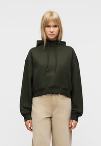 Γυναίκα με ίσια ξανθά μαλλιά φορώντας ένα σκουροπράσινο κοντό φούτερ με κουκούλα και μπεζ παντελόνι ψηλής μέσης σε απλό φωτεινό φόντο.