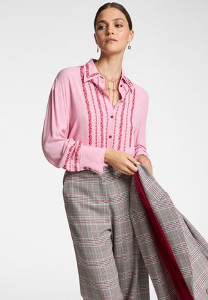 Blusa rosa con dettagli ricamati rossi, con bottone frontale e maniche lunghe, abbinata a pantaloni a quadri grigi con accenti rosa.