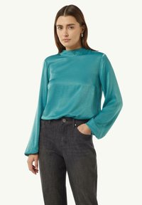 Türkise Bluse mit langen Ärmeln, hochgeschlossen und mit weichem, schimmerndem Material, kombiniert mit dunkelgrauen Jeans mit dezentem Muster.