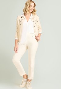 Beige vest met decoratieve bloemrijke borduursels over een wit shirt, gecombineerd met crèmekleurige broek en beige sneakers. Gladde gebreide textuur.