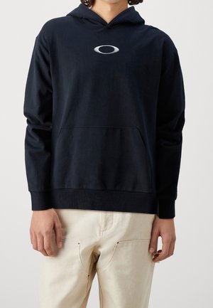 Person trägt schwarzen Hoodie mit weißem ovalem Logo auf der Brust und hellbeiger Hose vor schlichtem grauem Hintergrund.