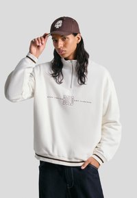 Svetlo kremast pulover s pol-zadrgo in ovratnikom, ki vsebuje besedilo "BOLD". Rebrasti manšeti z rjavimi akcenti, v kombinaciji s temnimi kavbojkami. Rjava kapa na glavi.