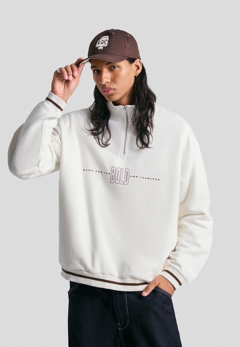 Svetlo kremast pulover s pol-zadrgo in ovratnikom, ki vsebuje besedilo "BOLD". Rebrasti manšeti z rjavimi akcenti, v kombinaciji s temnimi kavbojkami. Rjava kapa na glavi.