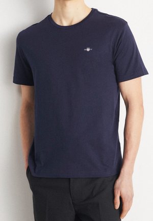 Man wearing a plain navy blue short-sleeve T-shirt with a small white logo on the left chest, paired with black pants.