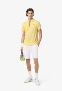 Polo giallo con zip, motivo diagonale, pantaloni corti bianchi e sneakers bianche. Tiene una borsa a rete con palline da tennis.