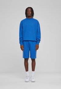 Felpa blu con maniche lunghe e pantaloni corti abbinati; entrambi gli articoli in morbido tessuto. Sneakers bianche e calzini completano l'outfit.